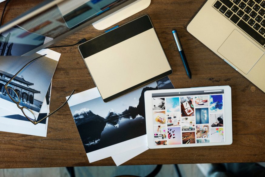 Afbeelding: Bovenaanzicht van een houten werkplek met een laptop, monitor en foto-prints ter illustratie van asset management bij fotografie