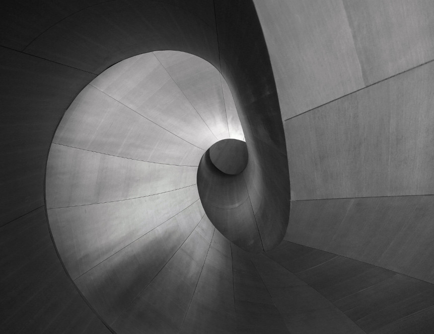 Afbeelding: Abstracte zwart-wit opname van een perfecte spiraalvormige trap die naar een centraal lichtpunt leidt, wat ritme en geometrie benadrukt en een goede compositie geeft aan de architectuurfotografie