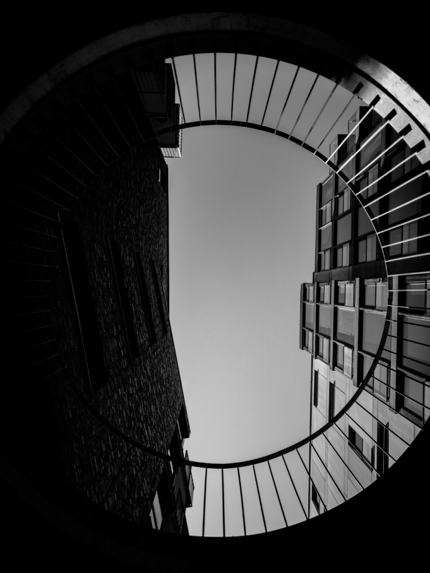Afbeelding: Artistieke zwart-wit foto vanuit een laag standpunt waarbij door een ronde betonnen opening omhoog wordt gekeken naar moderne gebouwen met strakke verticale lijnen
