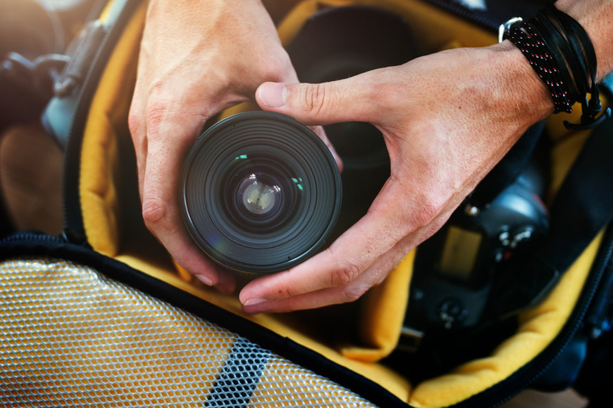 Afbeelding: handen van een fotograaf plaatsen voorzichtig een zwarte cameralens terug in de vakverdeling van een gevoerde, gele cameratas