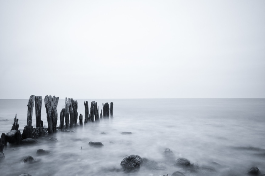 Afbeelding: Zwart-wit landschapsfotografie van een rustige zee met houten palen en een dromerige mist door lange sluitertijd