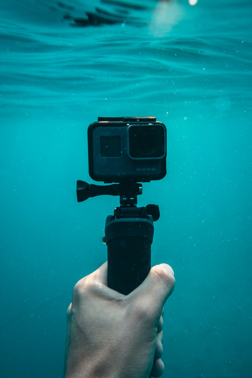 Afbeelding: hand houdt een zwarte GoPro actioncamera vast op een handgreep, net onder het wateroppervlak in helder blauw water