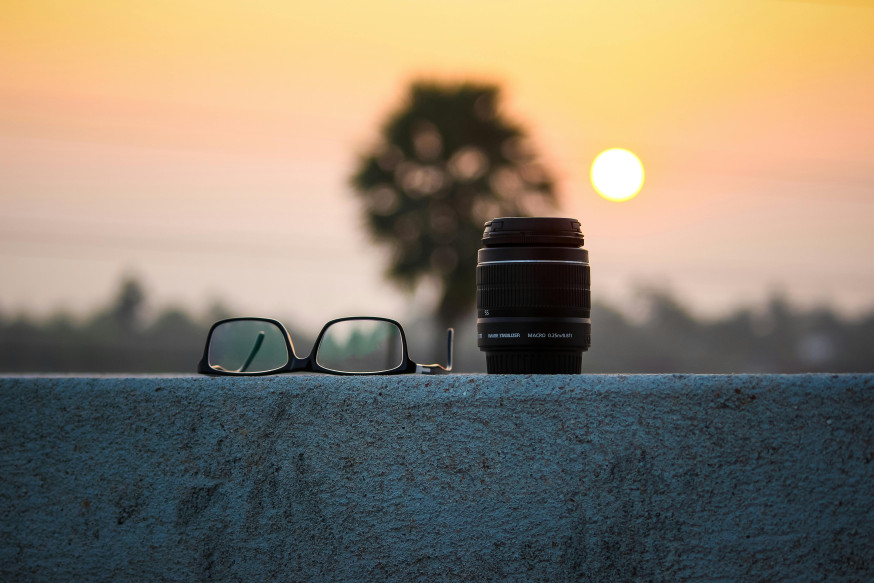 Afbeelding: Macro-objectief en bril op een muurtje met zonsondergang en zachte bokeh op de achtergrond