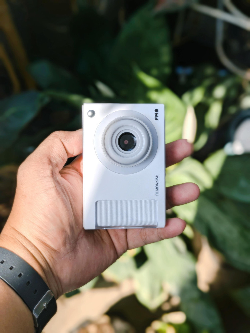 Afbeelding: Een hand houdt een minimalistische witte digitale camera vast tegen een groene achtergrond, symbool voor eenvoudige point-and-shoot fotografie